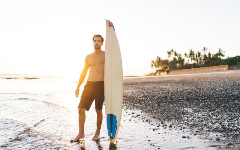 Handsome male dressed in black shorts posing at tropical coastline holding surfboard